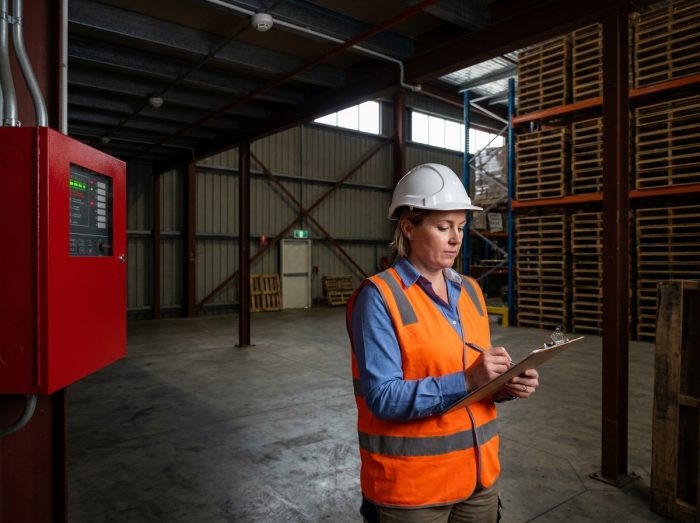 Consultante en sécurité incendie vérifiant un tableau d'alarme incendie dans un entrepôt industriel avec checklist de contrôle